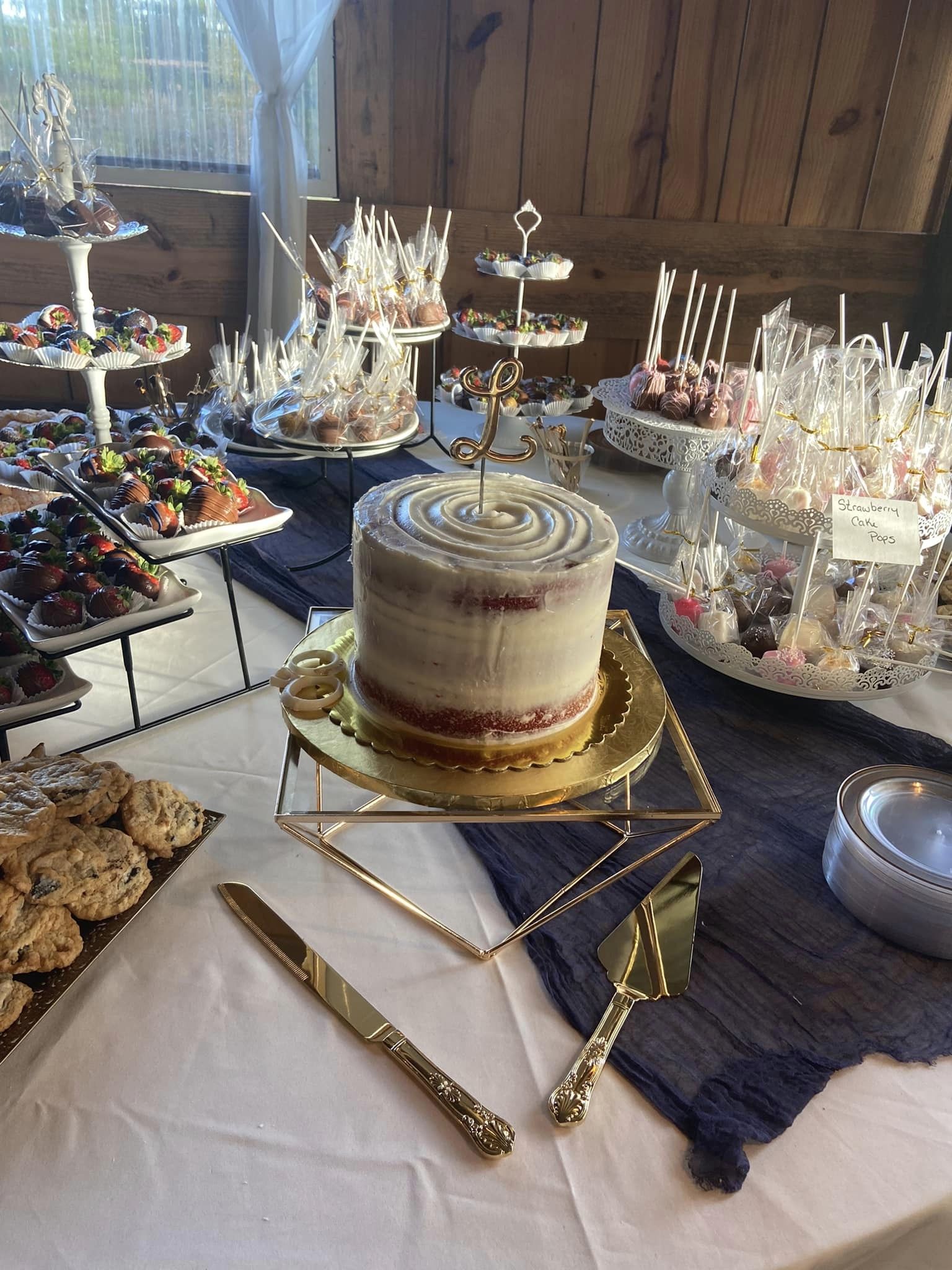 A dessert table by Rollover: a red velvet cake on a gold stand, cake pops, chocolate-dipped strawberries and cookies